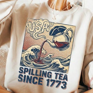 May include: Cream-colored sweatshirt featuring a graphic of a teapot pouring tea into ocean waves, with the text "USA" and "Spilling Tea Since 1773". The design is in a vintage style with red, blue, and cream colors.