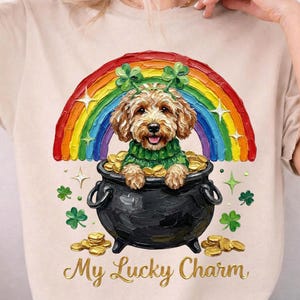 May include: A beige t-shirt featuring a colorful illustration of a dog in a pot of gold under a rainbow. The dog is wearing a green sweater and a shamrock headband. The text "My Lucky Charm" is below the image.