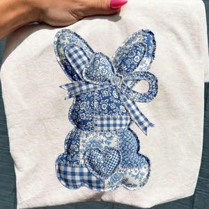 Könnte beinhalten: Ein weißes Handtuch mit einem blau-weißen Patchwork-Hasen-Design. Der Hase besteht aus verschiedenen gemusterten Stoffen, darunter Blumenmuster, Vichy-Karo und einfarbigem Blau. Der Hase hat einen herzförmigen Aufnäher und eine Schleife. Das Handtuch wird von einer Person gehalten.