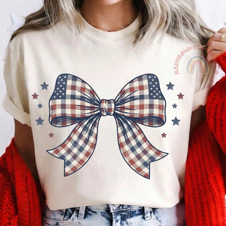 May include: Cream-colored t-shirt featuring a large bow graphic with a red, white, and blue plaid pattern. The bow design incorporates stars and stripes, with additional scattered stars. The shirt has short sleeves and a round neckline.