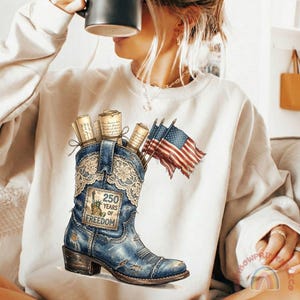 May include: Cream-colored sweatshirt featuring a distressed blue cowboy boot graphic. The boot is filled with rolled-up scrolls and American flags. The boot has a patch that reads "250 Years of Freedom".