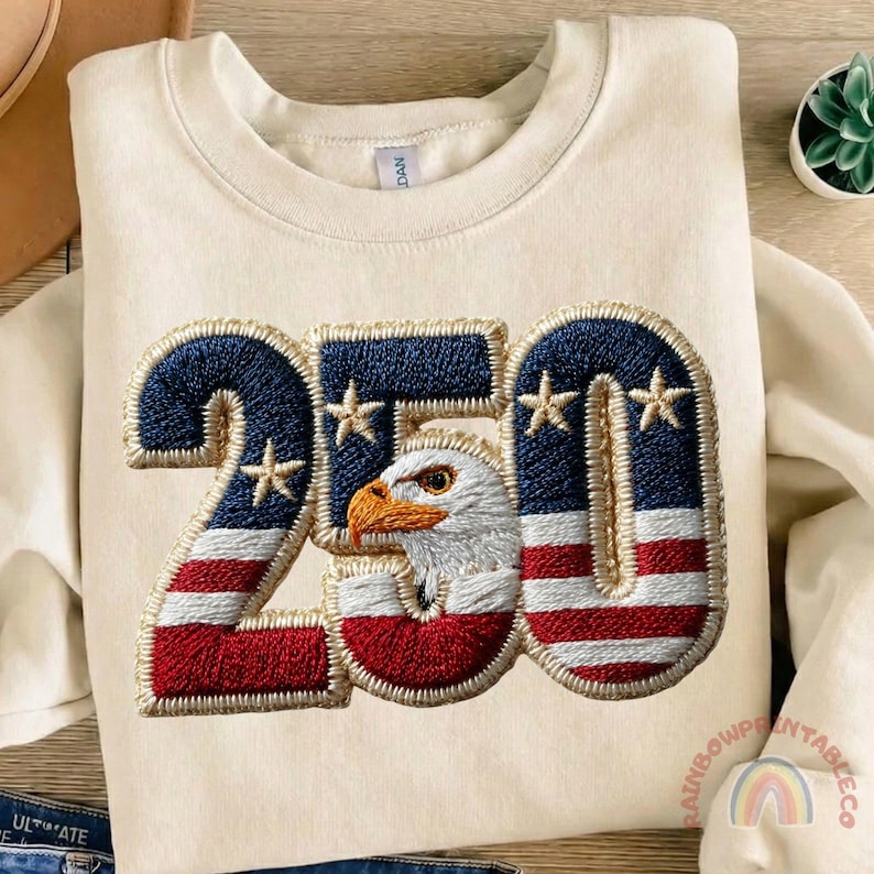 K&ouml;nnte beinhalten: Cremefarbenes Sweatshirt mit patriotischem Design. Das Design zeigt die Zahl "250" mit einem rot-wei&szlig;-blauen Muster der amerikanischen Flagge und einem Adlerkopf. Das Sweatshirt ist aus einem weichen Material gefertigt.