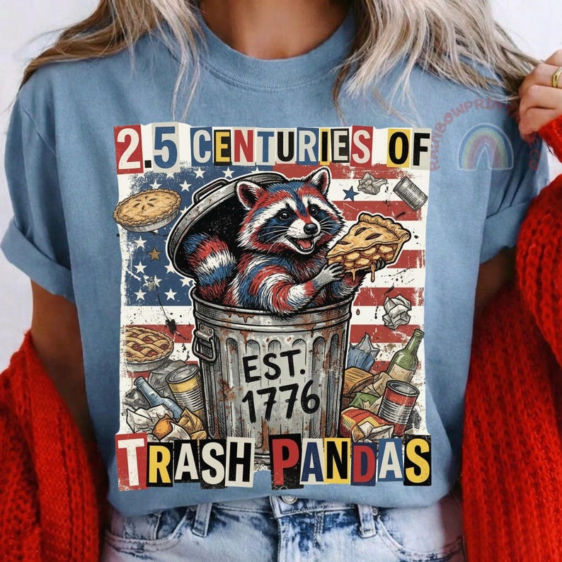 May include: Light blue t-shirt featuring a raccoon in a trash can, holding a slice of pie. The design includes the text "2.5 Centuries of Trash Pandas" and "Est. 1776" with an American flag background. Other pies and trash are also visible.