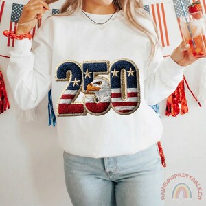 K&ouml;nnte beinhalten: Wei&szlig;es Sweatshirt mit patriotischem Design. Das Design zeigt die Zahl "250" in Rot, Wei&szlig; und Blau, mit einem Adler in der Mitte. Das Sweatshirt ist ein Rundhals-Stil.