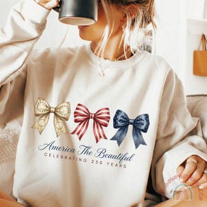 May include: Cream-colored sweatshirt featuring three watercolor bows in patriotic colors: tan with stars, red and white stripes, and navy with white stars. Below the bows is the text "America The Beautiful Celebrating 250 Years."