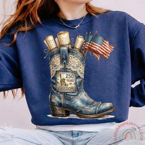 May include: A navy blue sweatshirt featuring a distressed denim-style cowboy boot with a lace overlay. The boot holds rolled scrolls and American flags. The boot has a patch that reads "250 Years of Freedom."