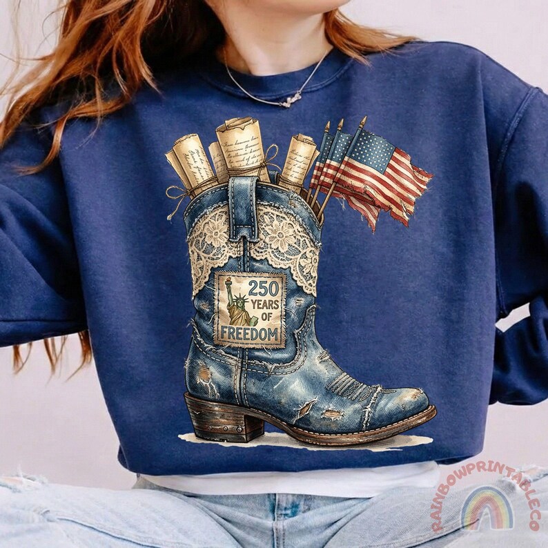 May include: A navy blue sweatshirt featuring a distressed denim-style cowboy boot with a lace overlay. The boot holds rolled scrolls and American flags. The boot has a patch that reads "250 Years of Freedom."