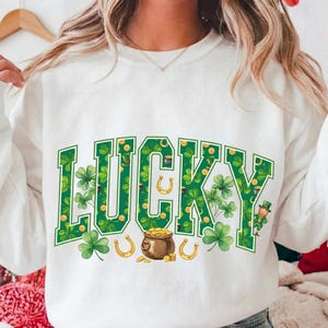 Puede incluir: Sudadera blanca con la palabra "LUCKY" en verde, decorada con tréboles, monedas de oro, herraduras y un duende. Las letras están rellenas con un patrón de trébol.