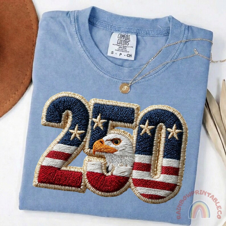 K&ouml;nnte beinhalten: Hellblaues Comfort Colors T-Shirt mit einem gestickten Design mit der Zahl "250" in Rot, Wei&szlig; und Blau, mit einem Adlerkopf in der Mitte. Eine goldene Halskette liegt &uuml;ber dem Shirt.