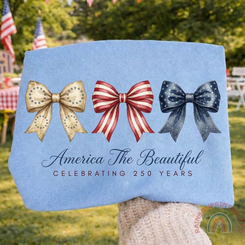 May include: A light blue towel featuring three decorative bows in patriotic colors: tan with stars, red and white stripes, and navy with stars. The text "America The Beautiful Celebrating 250 Years" is printed below the bows.