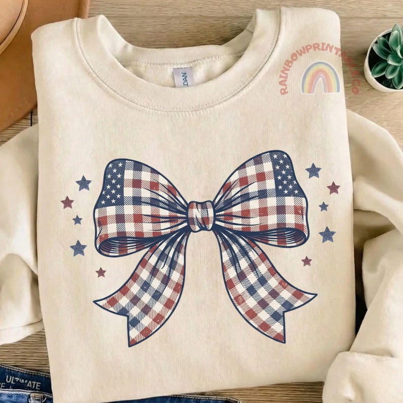 May include: Cream-colored sweatshirt featuring a large bow design in red, white, and blue plaid, with a section resembling the American flag. The design is surrounded by small blue and red stars. The text "RAINBOWPRINT" and a rainbow are visible.