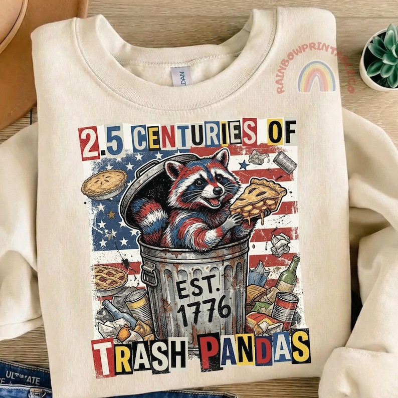 May include: Cream-colored sweatshirt with a graphic of a raccoon in a trash can, holding a slice of pie. The design includes the text "2.5 CENTURIES OF TRASH PANDAS" and "EST. 1776" with an American flag background.