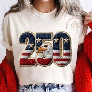 K&ouml;nnte beinhalten: Cremefarbenes T-Shirt mit patriotischem Design. Die Zahl "250" ist mit einem blau-rot gestreiften Muster bestickt, das Sterne und einen Adlerkopf enth&auml;lt. Der Text "RAINBOWPRINTS" und eine Regenbogen-Grafik sind auf dem Shirt zu sehen.