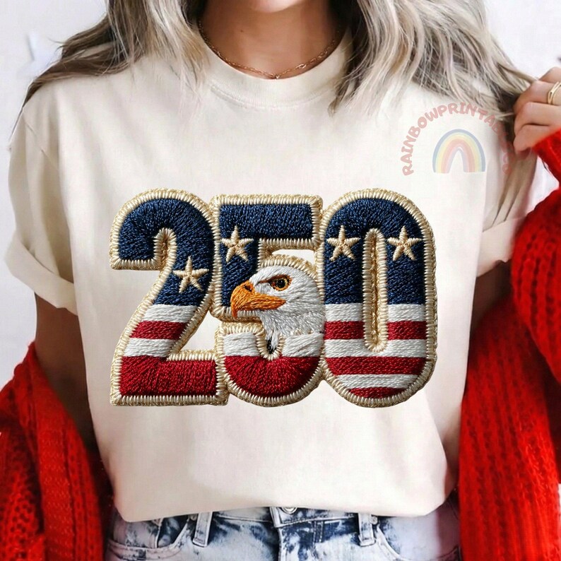K&ouml;nnte beinhalten: Cremefarbenes T-Shirt mit patriotischem Design. Die Zahl "250" ist mit einem blau-rot gestreiften Muster bestickt, das Sterne und einen Adlerkopf enth&auml;lt. Der Text "RAINBOWPRINTS" und eine Regenbogen-Grafik sind auf dem Shirt zu sehen.
