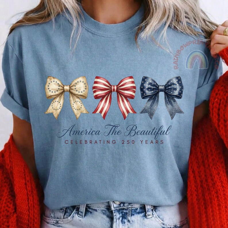 May include: Light blue t-shirt featuring three decorative bows in patriotic colors: gold with stars, red and white stripes, and navy with stars. The text "America The Beautiful" and "Celebrating 250 Years" are below the bows.