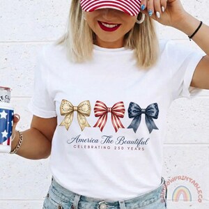 May include: White t-shirt featuring three decorative bows in gold, red and blue. The red bow has white stripes. The text "America The Beautiful CELEBRATING 250 YEARS" is printed below the bows.