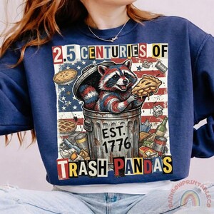 May include: Navy blue sweatshirt with a graphic of a raccoon in a trash can, holding pie. The design includes the text "2.5 Centuries of Trash Pandas" and "Est. 1776" with an American flag background.