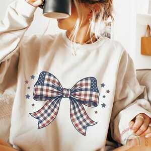 May include: Cream-colored sweatshirt featuring a large bow design with a plaid pattern in red, white, and blue, reminiscent of the American flag. The bow is surrounded by small blue and red stars, creating a patriotic theme.