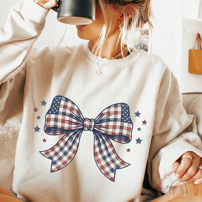 May include: Cream-colored sweatshirt featuring a large bow design with a plaid pattern in red, white, and blue, reminiscent of the American flag. The bow is surrounded by small blue and red stars, creating a patriotic theme.