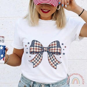 May include: White t-shirt featuring a large bow design with a red, white, and blue plaid pattern, surrounded by small stars. The bow design is centered on the shirt. The shirt is a casual style.
