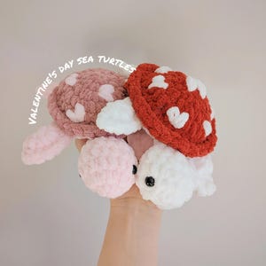 May include: Three crocheted sea turtle plushies in pink, red, and white. Each turtle has a heart-shaped pattern on its shell. The text "Valentine's Day Sea Turtles" is written in white on a pink background.