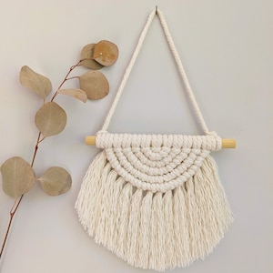 May include: A white macrame wall hanging with a wooden dowel and fringe. The hanging is in the shape of a half circle.