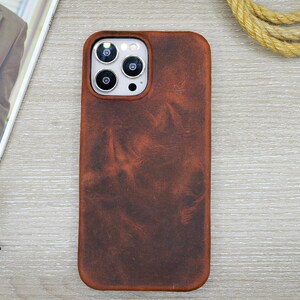 Könnte beinhalten: Eine braune Leder-Handyhülle mit einer strukturierten Oberfläche. Die Hülle hat einen Kameraausschnitt und ein glattes, abgerundetes Design. Die Handyhülle liegt auf einer hellen Holzmaserung, mit einer Zeitschrift, einer Brille und einem weißen kabellosen Kopfhörer-Etui im Hintergrund.