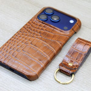 Pode incluir: Uma capa de smartphone castanha com um padrão texturado de pele de crocodilo e um porta-chaves de pele a condizer com um anel de metal dourado. A capa está colocada num smartphone azul escuro com três lentes de câmara visíveis.
