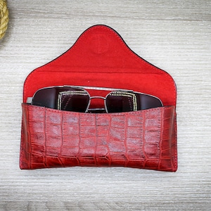 May include: A red leather sunglasses case with a crocodile pattern. The case is open and a pair of black sunglasses are inside.