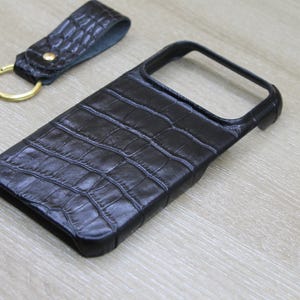 Puede incluir: Una funda de teléfono de color marrón oscuro con un patrón texturizado de piel de cocodrilo y un llavero de cuero a juego con un anillo de metal dorado. La funda tiene un gran recorte rectangular para la cámara y un teléfono blanco.