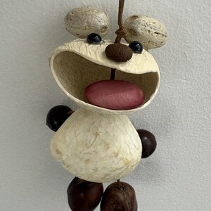 Può includere: Un ornamento fatto a mano a forma di orso sorridente. L'orso è realizzato con materiali naturali, con un corpo color crema, dettagli marrone scuro e una lingua rossa. È appeso a una corda marrone.