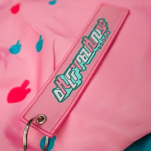 May include: Pink keychain tag with the text "DHRATHNY" in a teal and white design. The tag has a silver key ring and is set against a pink background with teal and red shapes.