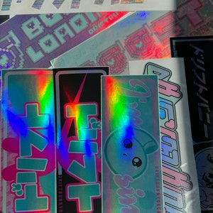 May include: A collection of holographic stickers with various designs and text. The stickers feature Japanese text, pixelated graphics, and the words "DRIFT BUNNY DECALS". Colors include pink, blue, and silver.