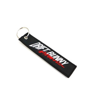 May include: Black rectangular keychain tag with the text "DRIFT BUNNY" in white and red lettering. The tag has a silver metal ring attached for easy attachment to keys or other items. The background is white.