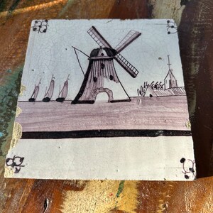 Op de afbeelding: Vierkante keramische tegel met een handgeschilderde scène van een windmolen, zeilboten en een kerk. Het ontwerp is in zwart, paars en wit, met decoratieve hoekaccenten. De tegel heeft een verouderde uitstraling.