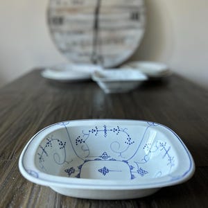 Op de afbeelding: Wit keramisch serveerschaaltje met een blauw bloemmotief. De rechthoekige schaal heeft afgeronde hoeken en een blauwe rand. Andere witte schalen en een decoratieve klok staan op de achtergrond. De schaal staat op een donkere houten tafel.