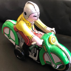 Op de afbeelding: Vintage blikken speelgoedmotor met een rijder. De motor is voornamelijk groen met witte en rode accenten. De rijder draagt een gele jas, een rode broek en een zilveren helm. Het speelgoed heeft zwarte wielen en staat tegen een donkere achtergrond.