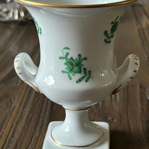 Op de afbeelding: Witte porseleinen vaas met een vierkante voet en twee handvatten. De vaas heeft groene bloemmotieven en gouden randen langs de rand, handvatten en voet. De vaas heeft een klassiek, elegant ontwerp.