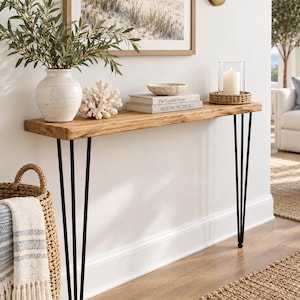 May include: A long, narrow console table with a natural wood grain top and four slender black metal hairpin legs. The table surface holds a white ceramic vase with green branches, a white coral sculpture, two stacked books, and a candle in a glass.