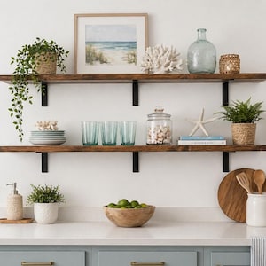 May include: A kitchen scene with two dark wooden shelves holding decorative items. Items include a framed beach print, glass bottles, coral, plants in woven baskets, plates, glasses, and a jar of shells. A wooden bowl of limes sits on the counter.