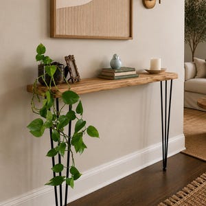 May include: A narrow console table featuring a natural light wood slab top supported by four slender black metal hairpin legs. The table holds a potted green plant, a small ornate picture frame, a stack of two books, a bird figurine, and a candle.