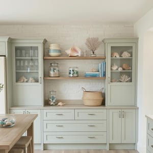 May include: A light-filled kitchen with a coastal theme. Features include light green cabinetry, a wooden table with stools, and decorative seashells. A glass pendant light hangs above the table. The room has a light wood floor and a white brick wall.