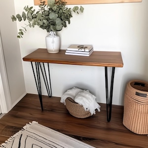 May include: A wooden console table with black hairpin legs, a white vase with green leafy branches, a stack of books, and a woven basket.