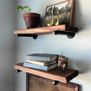 May include: Two wooden shelves with black metal brackets, mounted on a white wall. The top shelf has a framed picture with two gold bird figurines. The bottom shelf has a stack of books and a small wooden ship figurine.