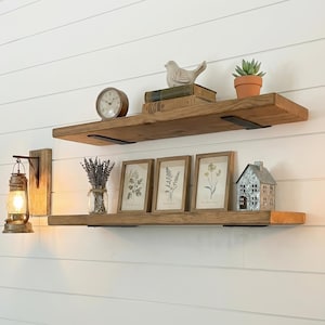 May include: Two rustic wooden shelves mounted on a white wall, adorned with decorative items. The top shelf holds a clock, books, a bird figurine, and a potted succulent. The bottom shelf displays framed botanical prints, a metal house, and a lantern.