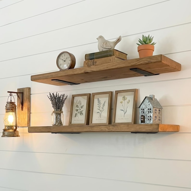 May include: Two rustic wooden shelves mounted on a white wall, adorned with decorative items. The top shelf holds a clock, books, a bird figurine, and a potted succulent. The bottom shelf displays framed botanical prints, a metal house, and a lantern.