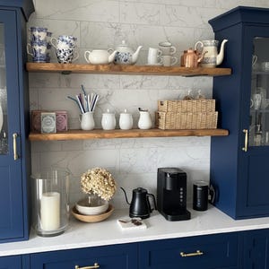 May include: A kitchen display with shelves holding teacups, teapots, and a coffee maker. The shelves are made of wood and are mounted on a white marble tile wall. The cabinets are navy blue. There is a glass candle holder and a small basket.