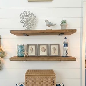 May include: Coastal-themed shelf decor featuring wooden shelves with various items. Includes framed seashell prints, a blue mason jar, a starfish, a decorative lighthouse, a bird figurine, and a small potted plant. The background is a white shiplap wall.