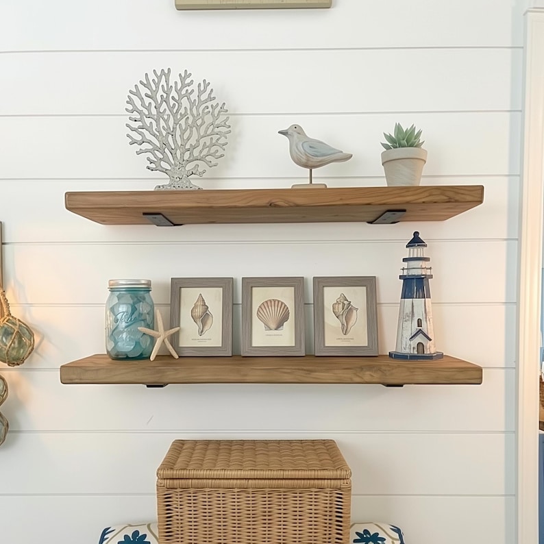 May include: Coastal-themed shelf decor featuring wooden shelves with various items. Includes framed seashell prints, a blue mason jar, a starfish, a decorative lighthouse, a bird figurine, and a small potted plant. The background is a white shiplap wall.