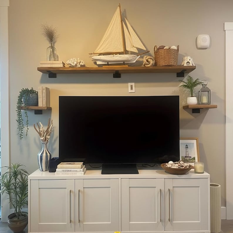 May include: A coastal-themed living room with a white cabinet, a large black television, and floating shelves. The shelves display a model sailboat, decorative items, and plants. A framed lighthouse picture and a candle are on the cabinet.
